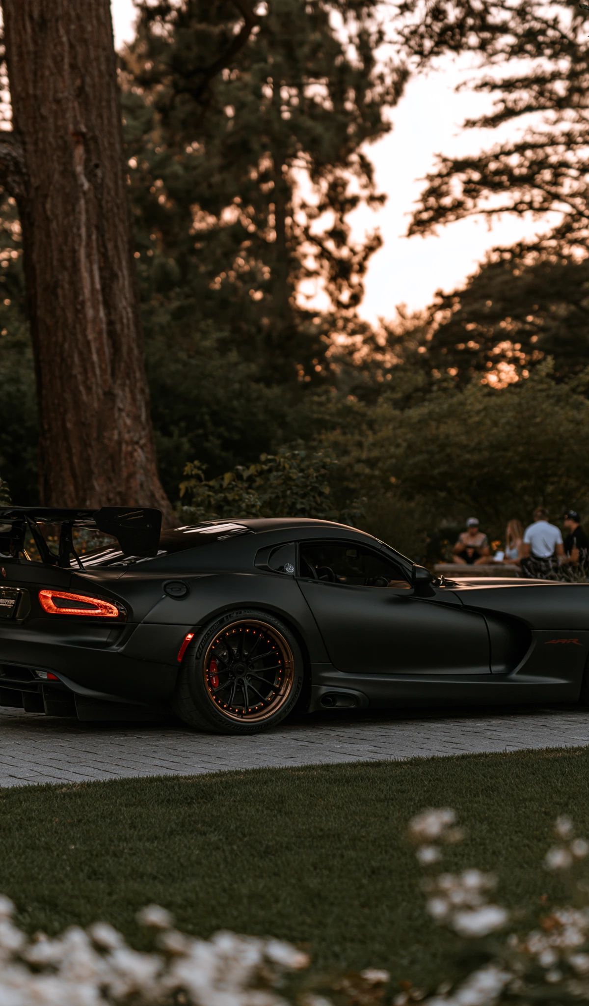 A matte black sports car with red-accented wheels parked on a paved area surrounded by tall redwood trees at sunset