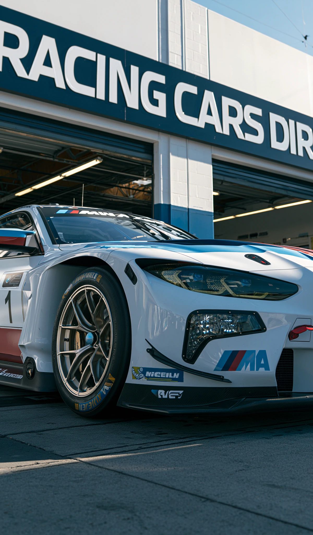 A white, blue, and red BMW M racing car with aerodynamic bodywork parked in front of a BMW Racing Cars Direct facility storefront