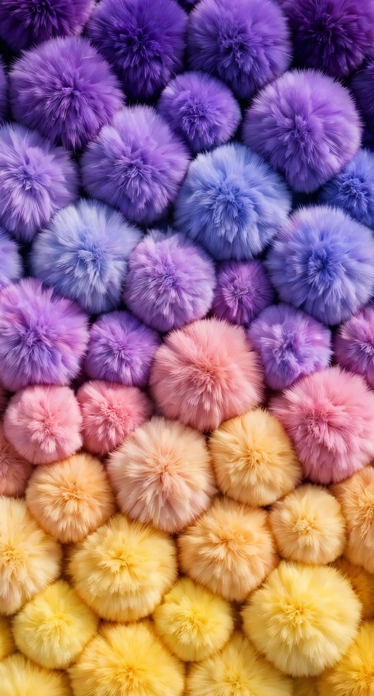 Overhead view of numerous soft, fuzzy pompoms arranged in a gradient from yellow at the bottom through pink in the middle to purple and blue at the top